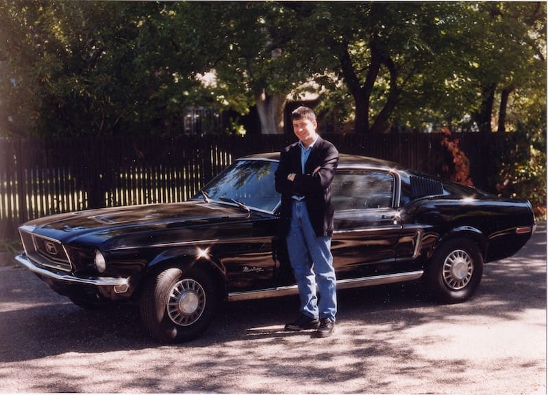 Teenage boy in jeans and a sport coat standing outside in front of a black 1968 Mustang fastback