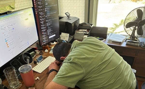Man in green shirt with his head on a cluttered desk, two monitors showing open screens.